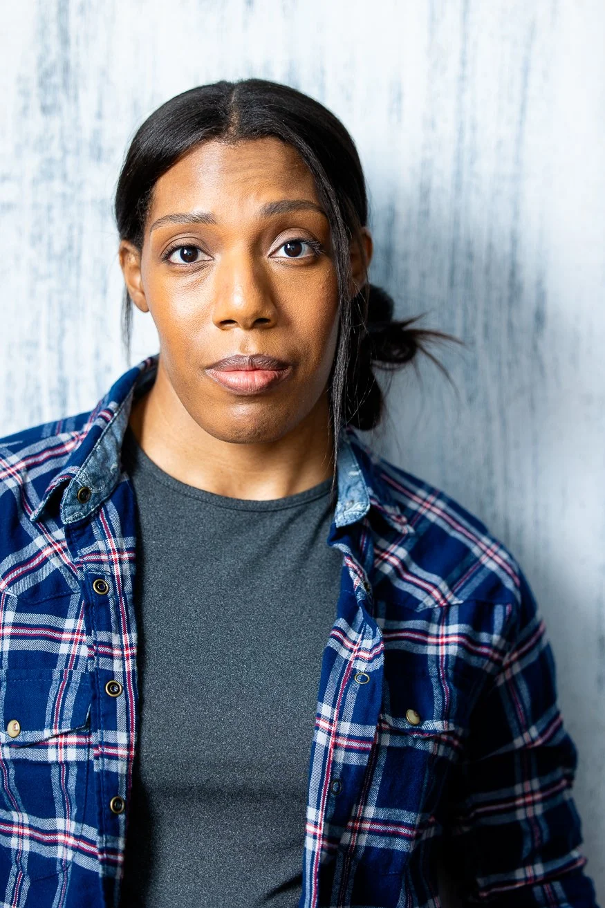 An African American woman with dark-brown hair styled in a side bun, wearing a blue plaid shirt over a gray t-shirt, standing against a light-colored wooden wall.