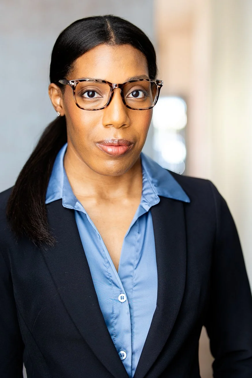 A professional African American woman with dark-brown hair styled in a ponytail, wearing glasses and a black blazer over a blue collared shirt, standing in a well-lit indoor setting.