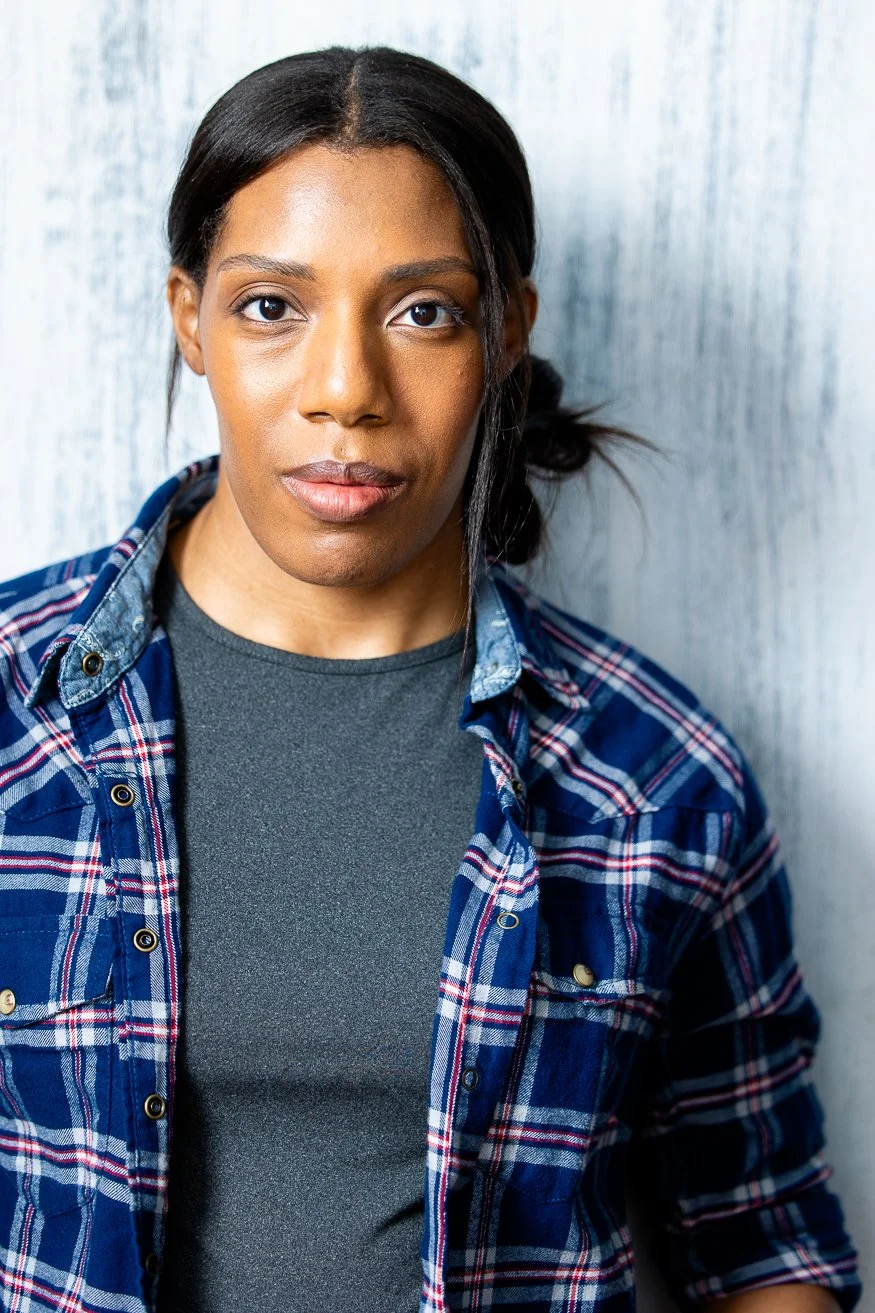 An African American woman with dark-brown hair styled in a side bun, wearing a blue plaid shirt over a gray t-shirt, standing against a light-colored wooden wall.