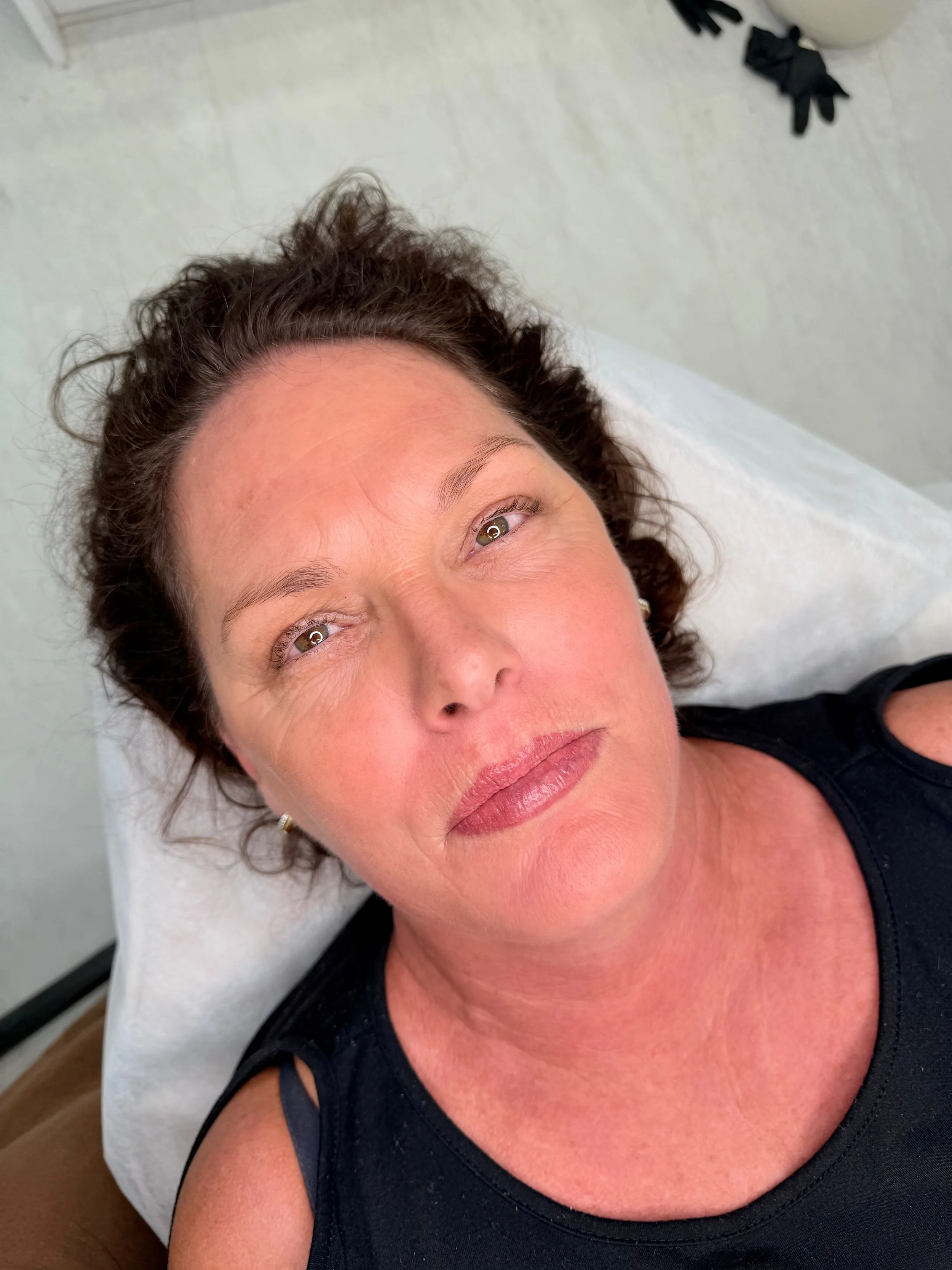 Close-up of a middle-aged woman with brown curly hair, lying on a white pillow, wearing a black sleeveless top, looking directly at the camera with a neutral expression.