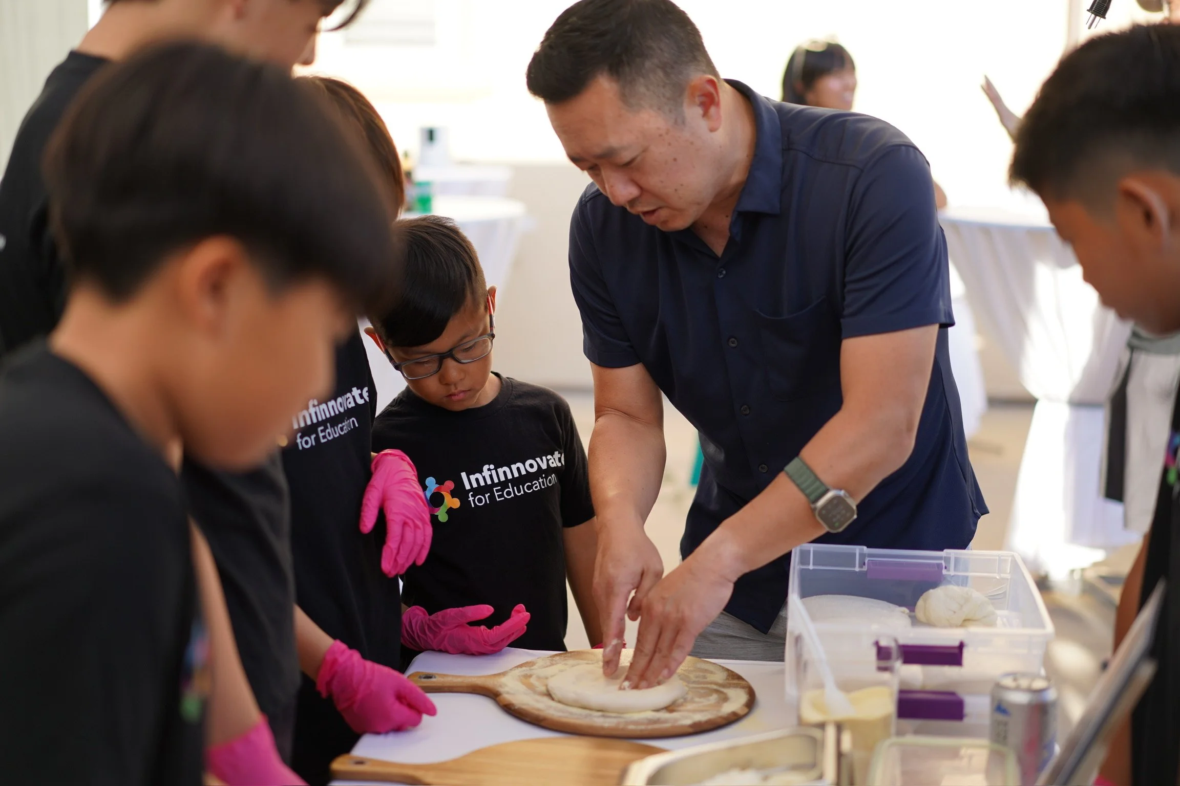 A group of children and an adult making pizza together at a table in a bright room, with some children wearing black shirts that say "Infnnovate for Education." The adult is pressing dough with his hands while the children observe closely.