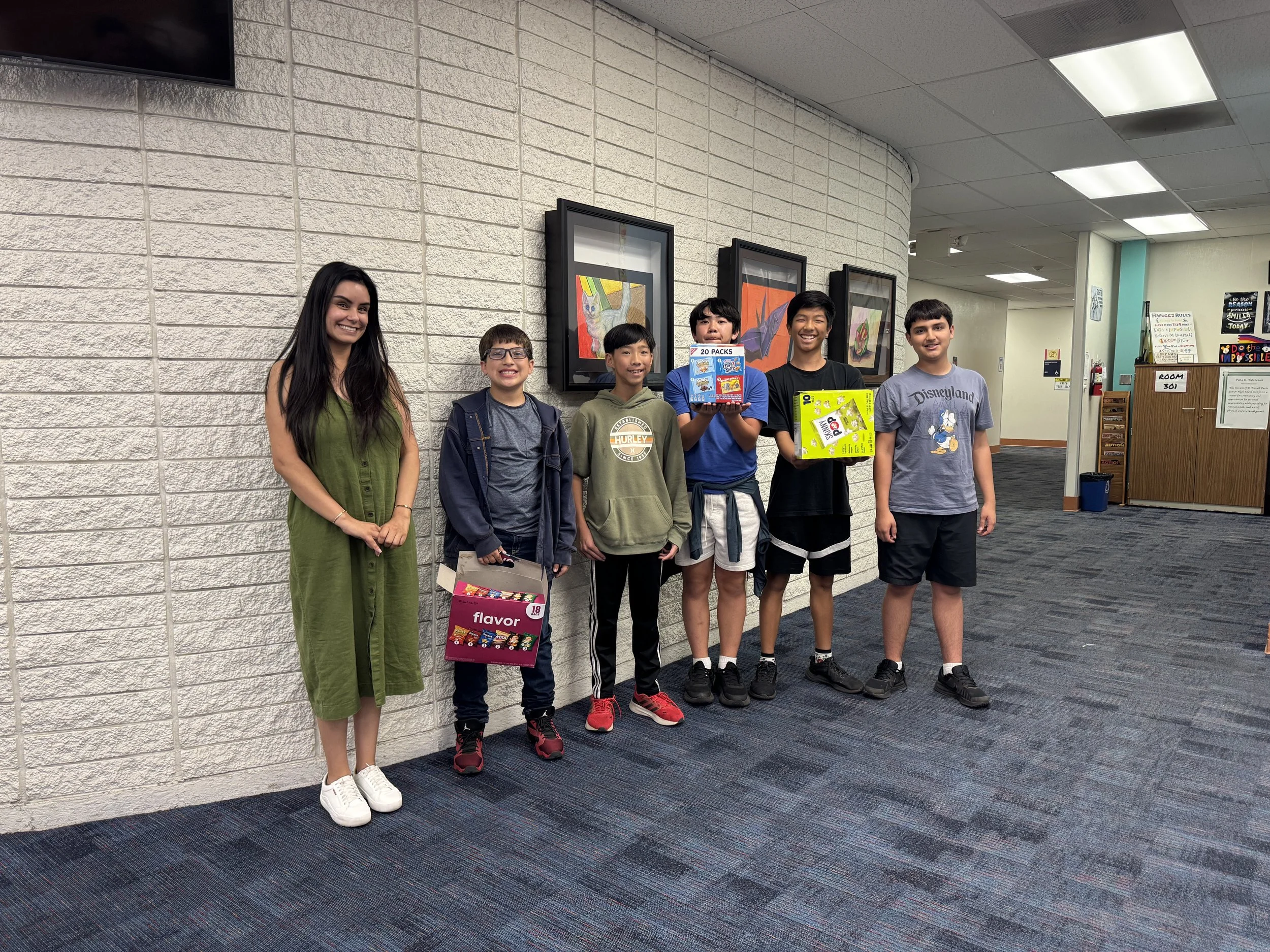 A group of six people, including a woman and five boys, standing indoors against a white brick wall with framed artwork, smiling at the camera. Some boys are holding boxes of snacks.