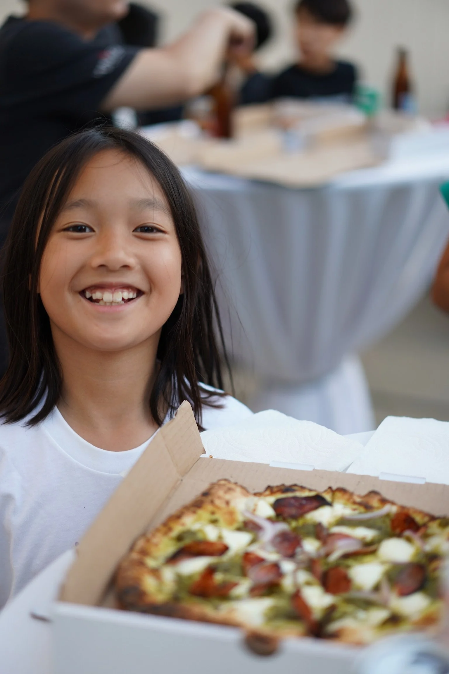 A young girl with long dark hair smiling and holding a pizza box with a pizza inside, in a room with people in the background.