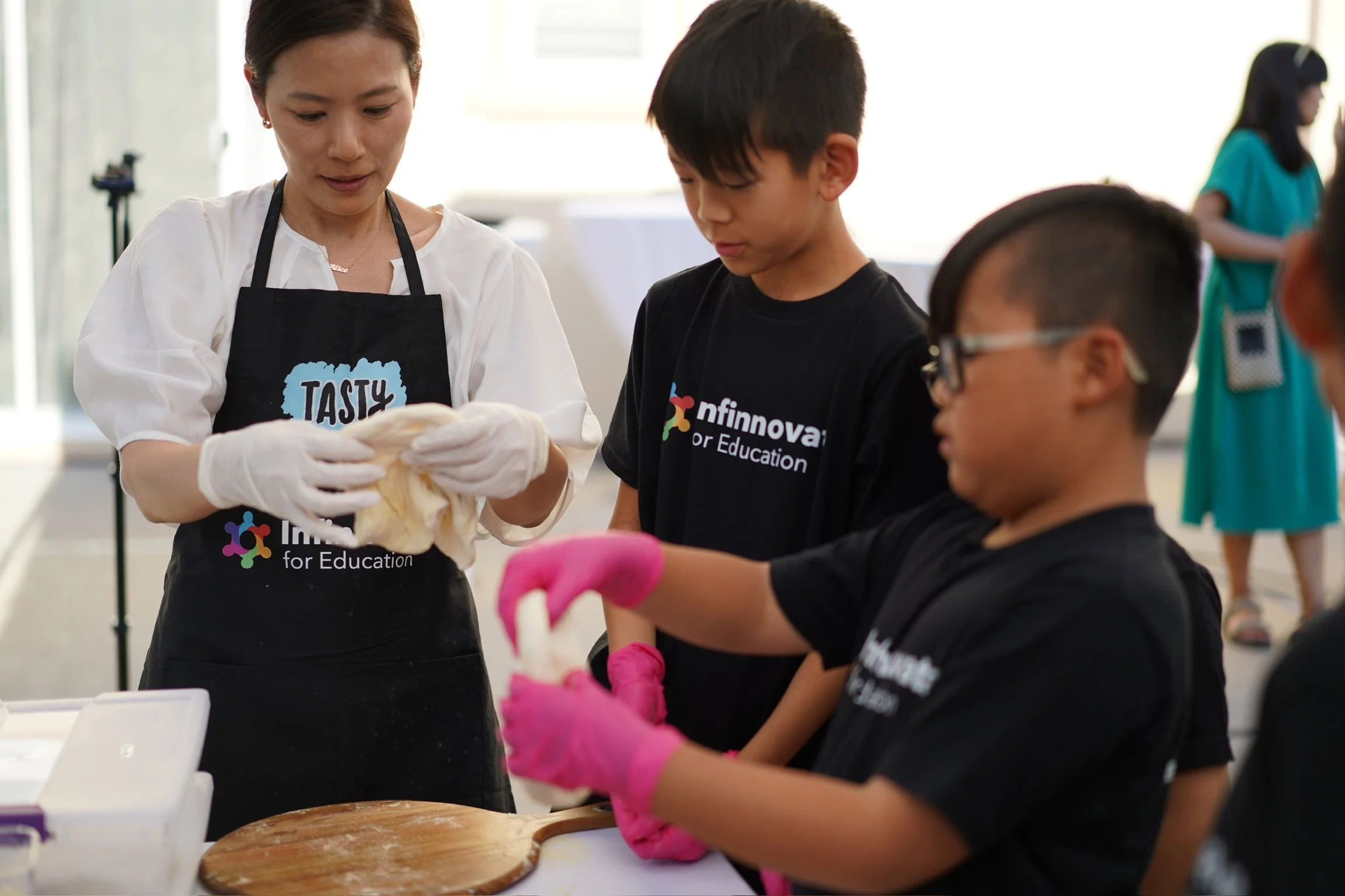 A woman instructs a group of children in a cooking or baking class, with all participants wearing black aprons with the 'nfinnovate for Education' logo. The woman is holding a dough or bread, and the children are wearing pink gloves, with one child in glasses looking at the activity. Other people are visible in the background.