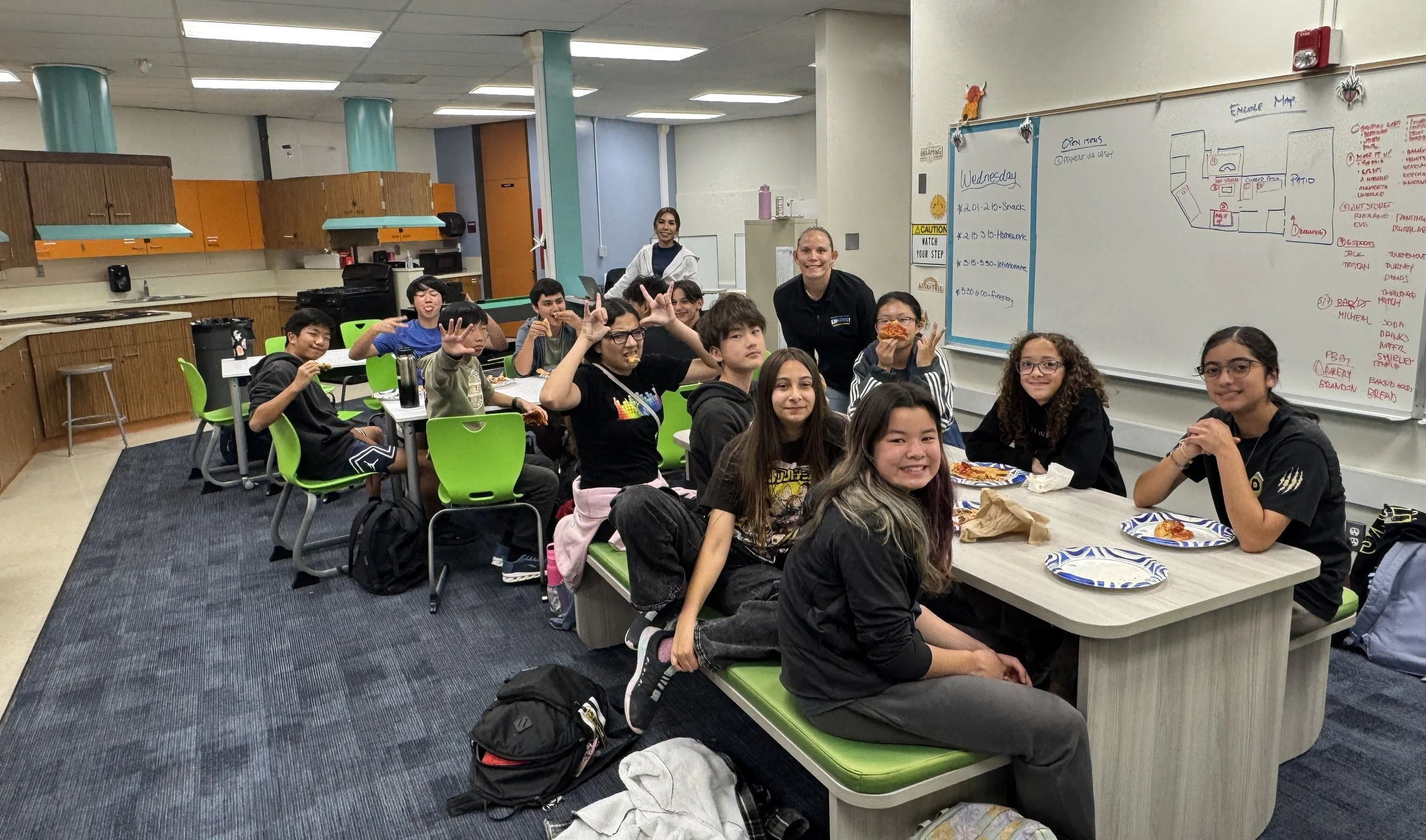 A group of students sitting around a table in a classroom, smiling, some holding pizza slices, with a teacher or facilitator standing behind them, and a whiteboard with notes and a diagram in the background.