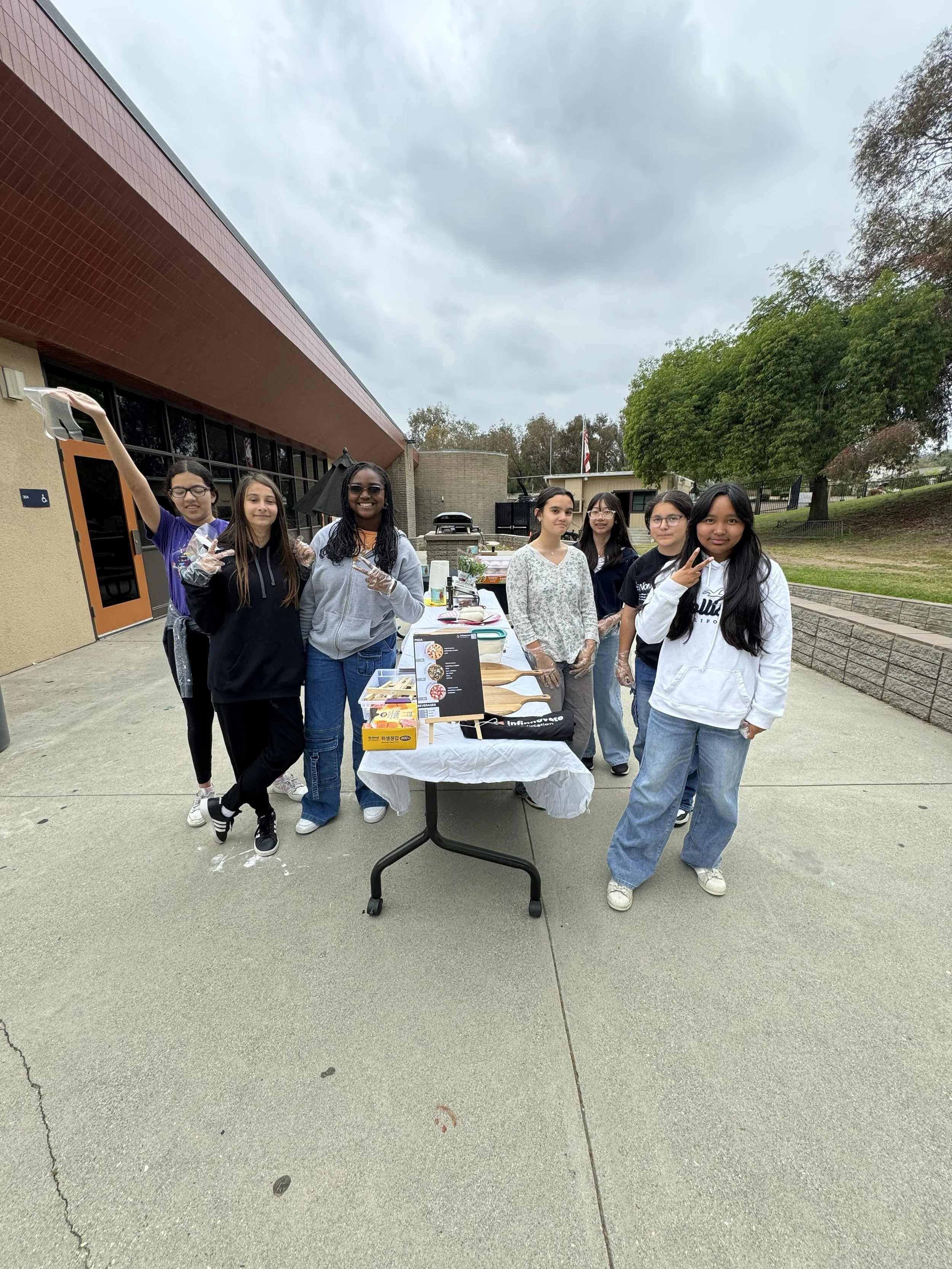 A group of seven young women standing behind a table outside on a cloudy day, with greenery and a building in the background.