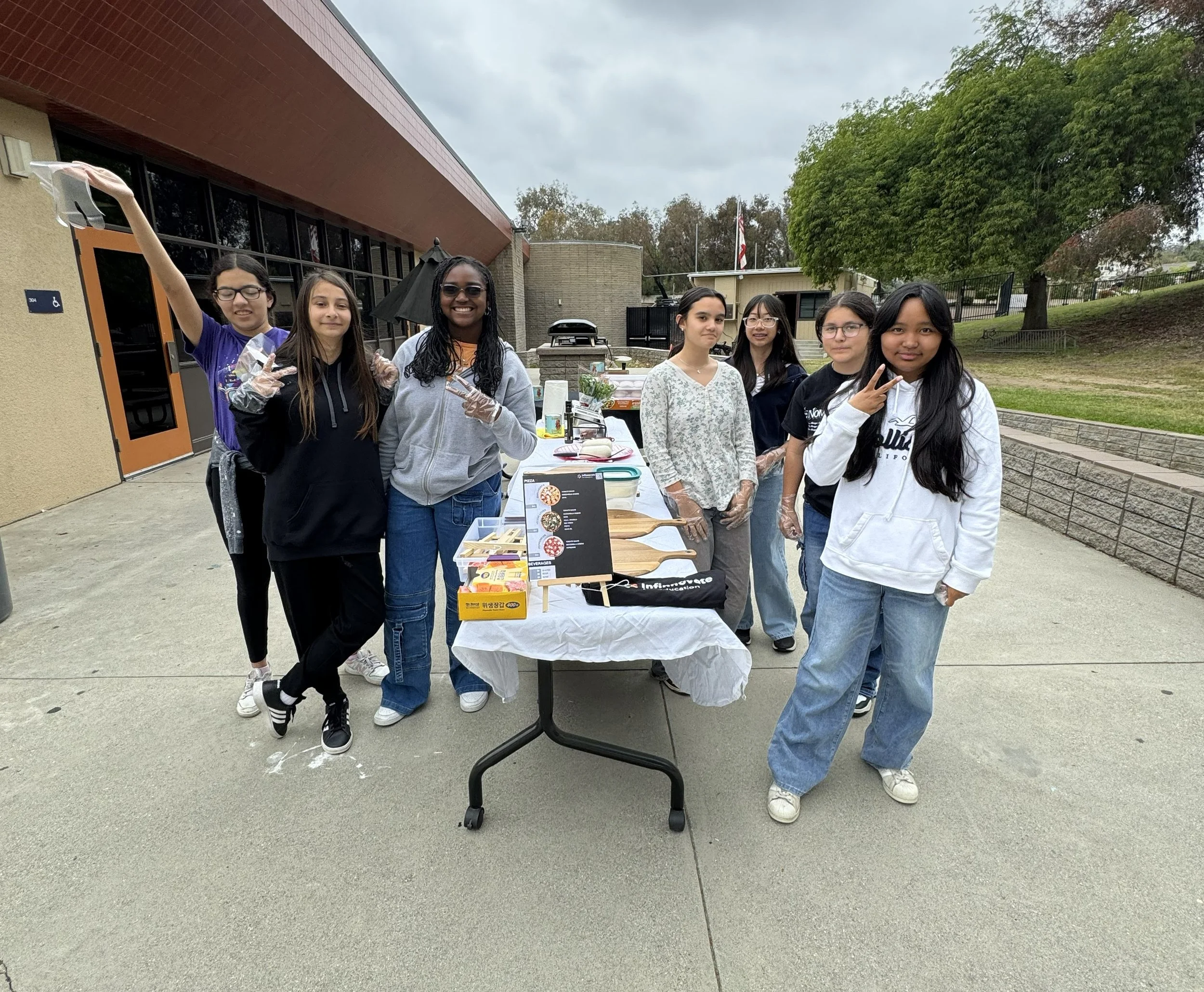 A group of seven diverse young girls standing outdoors behind a table with pizza and snacks, some wearing gloves, smiling and making peace signs. Overcast sky, trees, and a building in the background.