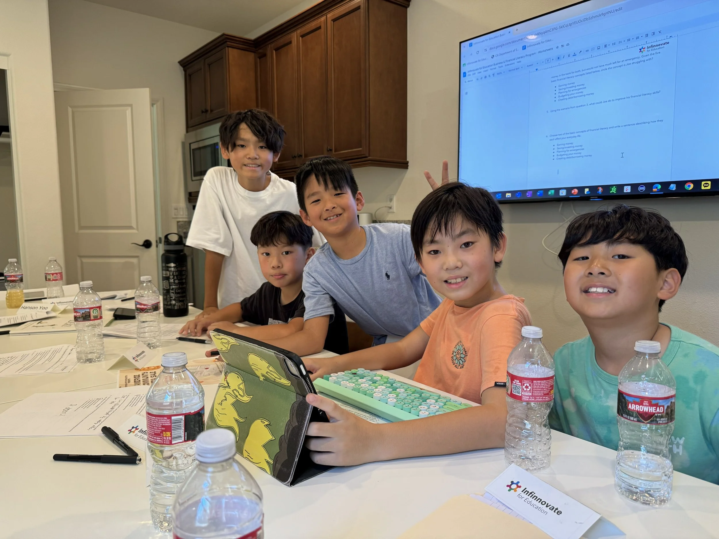 Group of five children sitting at a table during a classroom activity, with water bottles, papers, and a learning tool in front of them. A large screen displays educational content in the background.