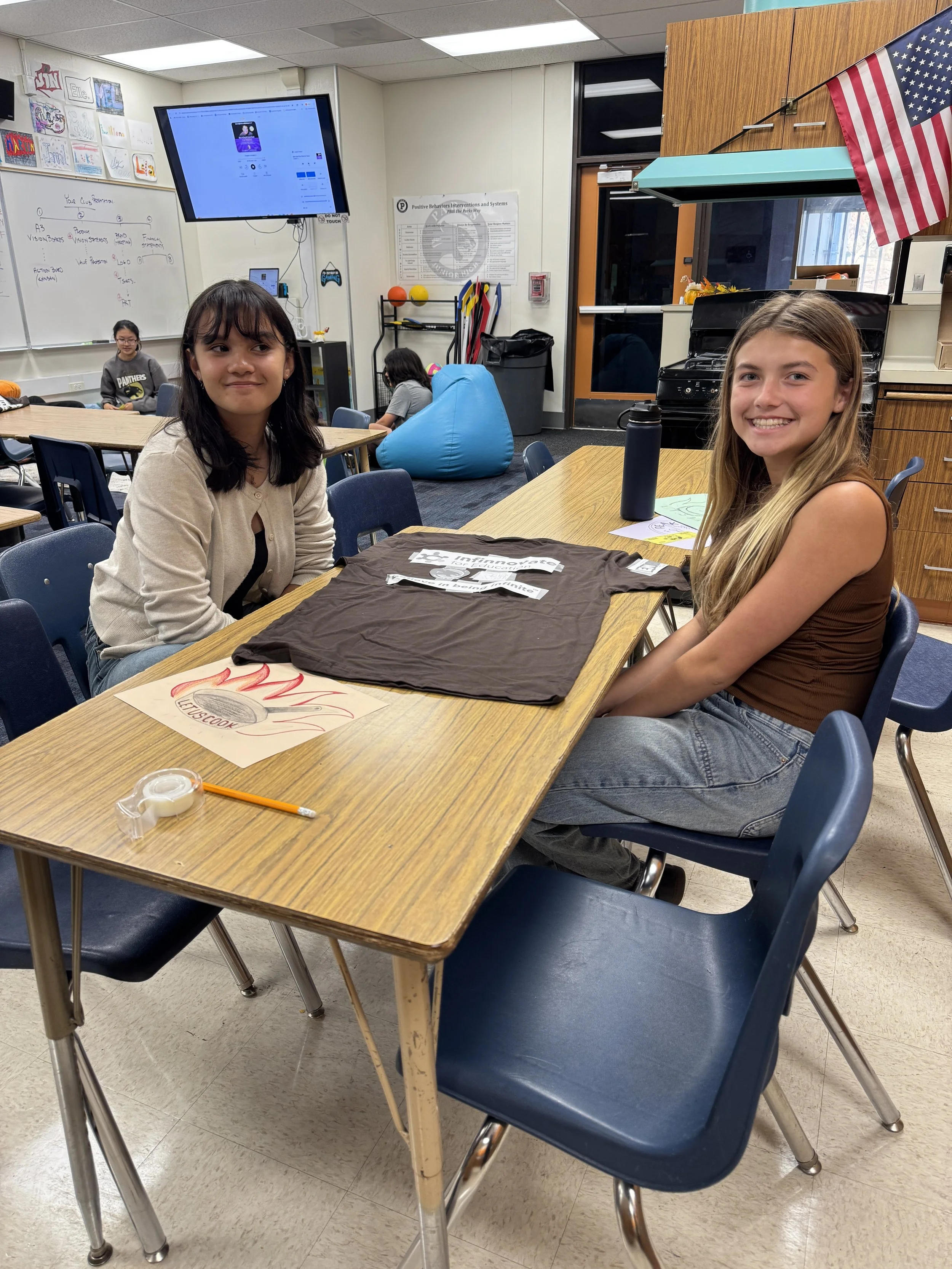 A classroom scene with students sitting at desks. Two girls are in the foreground, smiling and looking at the camera. One girl has dark hair and a beige cardigan, the other has long blonde hair and a brown top. There are other students in the background, artwork on the walls, a whiteboard, a large screen, and classroom equipment. An American flag is visible in the top right corner.