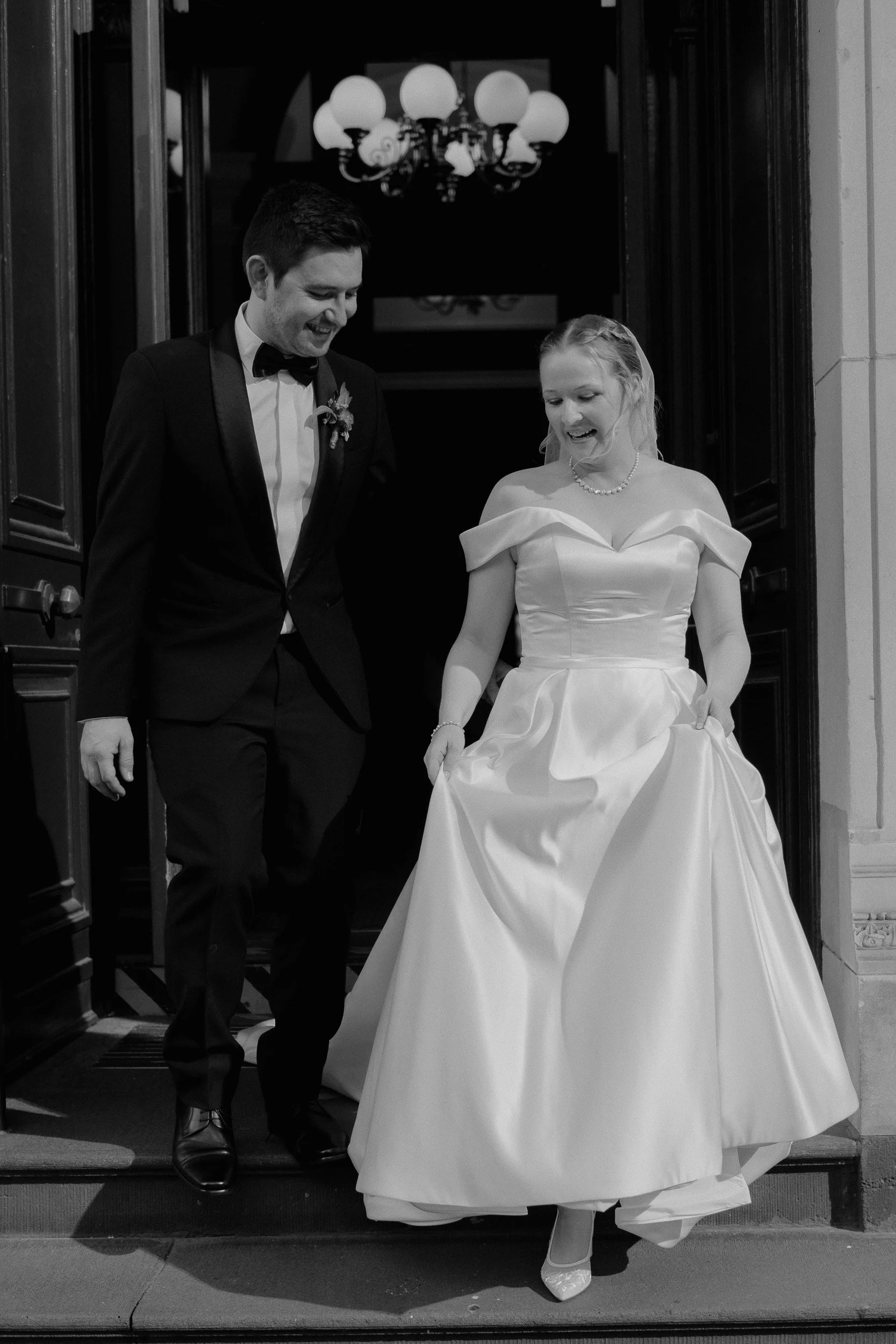 A black and white photo of a newlywed couple stepping out of a building, holding hands, with the bride lifting her gown. They are smiling, dressed in formal wedding attire, with the groom in a tuxedo and the bride in a wedding dress with a pearl necklace. A chandelier is visible in the background.