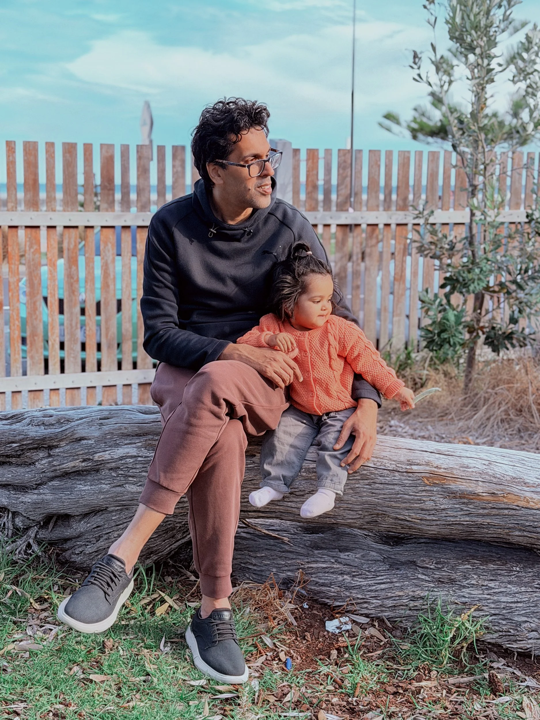 A man and a young girl sitting on a large fallen tree trunk outdoors, with a wooden fence, some plants, and a cloudy sky in the background.