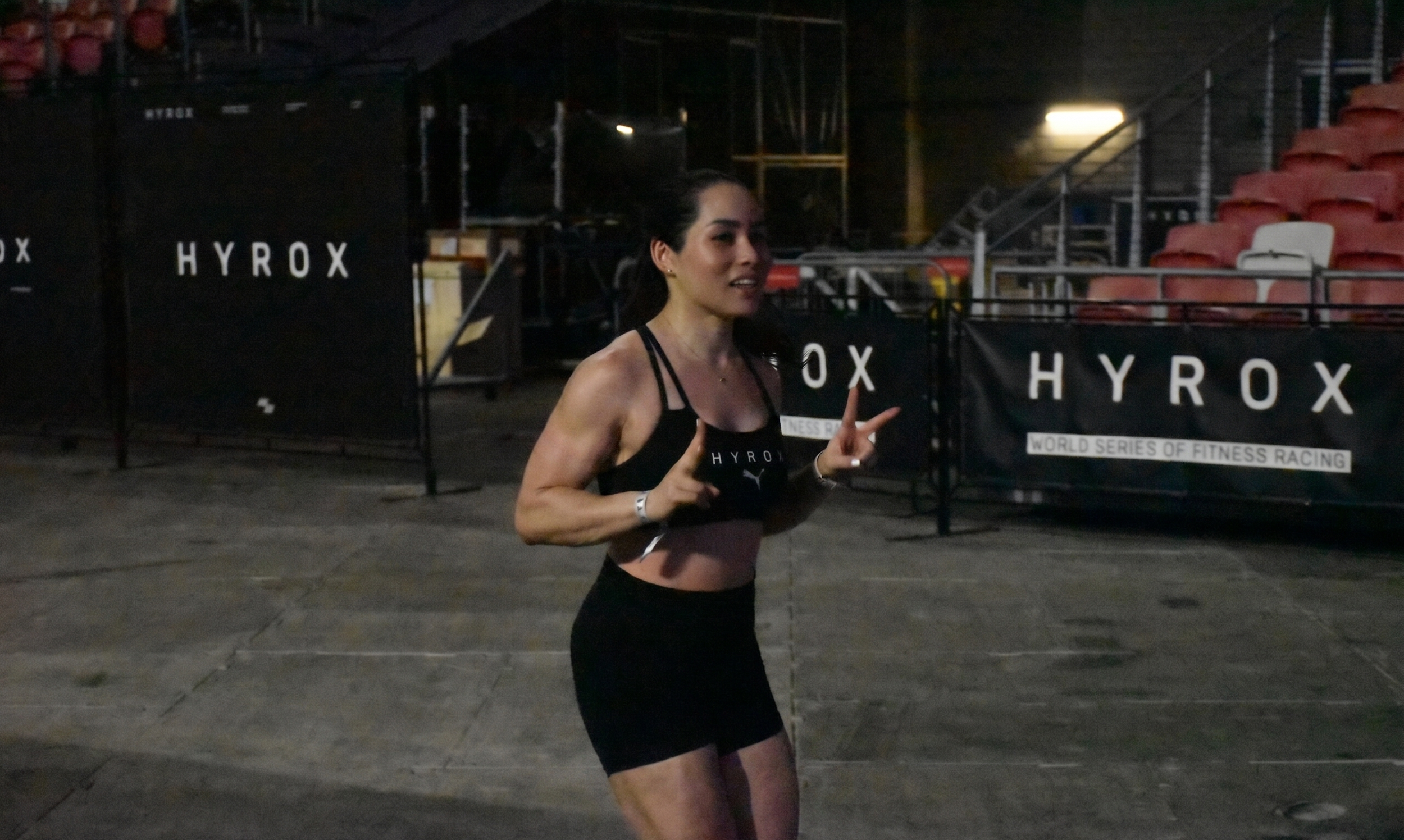 A woman running at a nighttime event, wearing a black sports bra and shorts with the brand Puma, near barriers with 'HYROX' branding, indicating a fitness race or marathon.