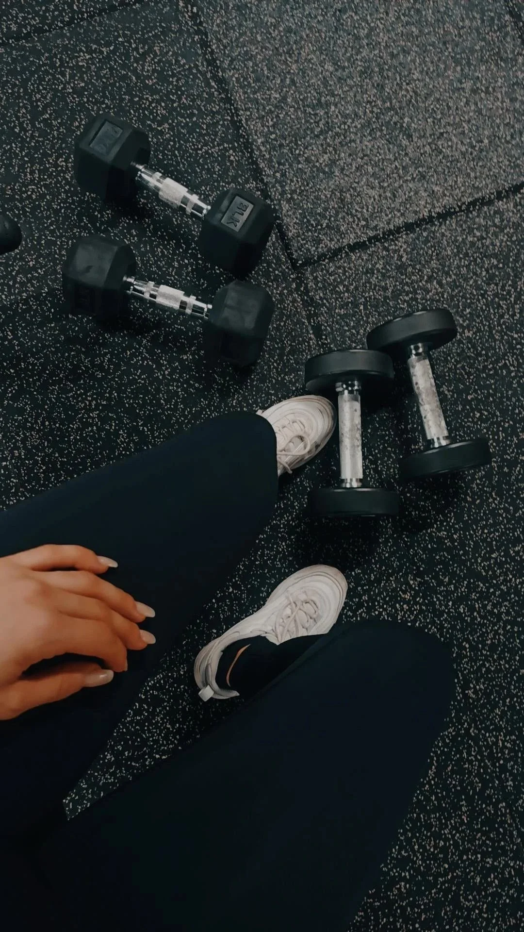 Gym workout area with two dumbbells, two kettlebells, a pair of white sneakers, and a person's hand resting on their leg, on a speckled rubberized gym floor.