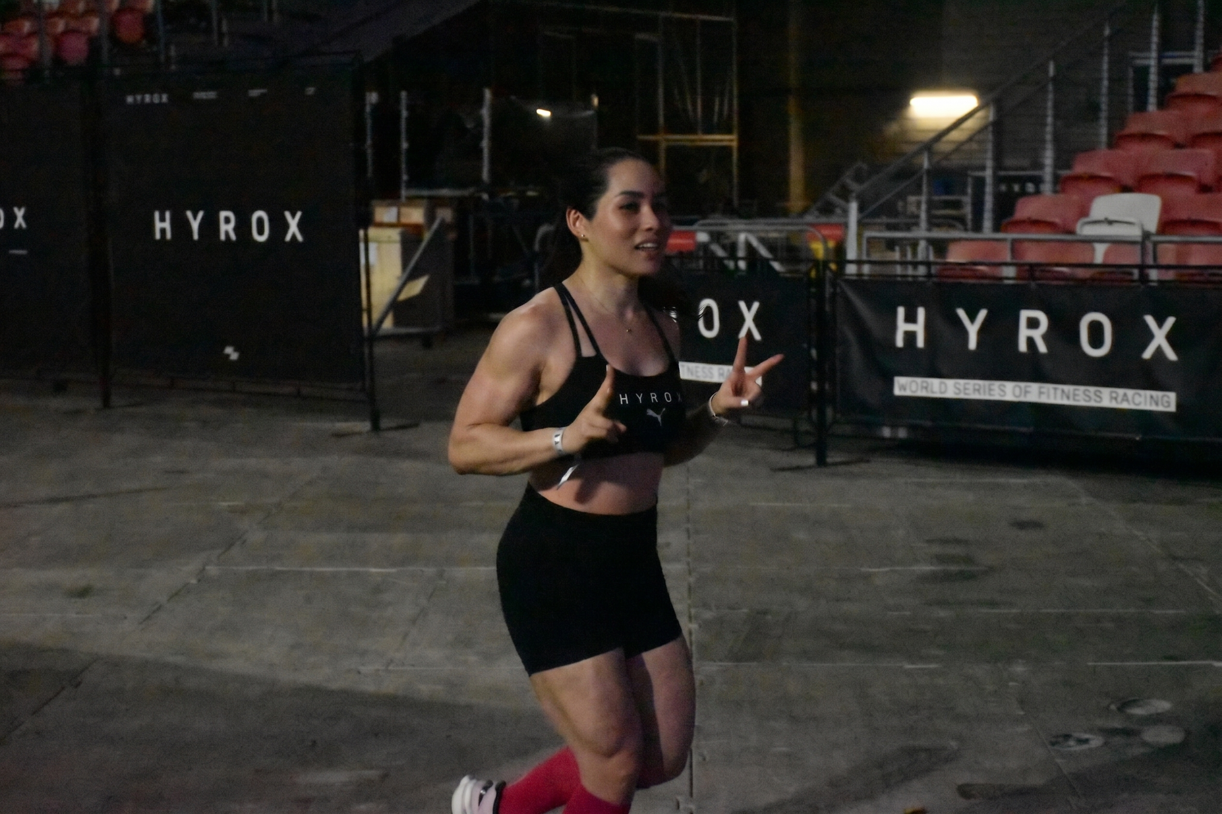 A woman running indoors at a fitness race event, wearing a black sports bra and black shorts with red compression socks, making peace signs with both hands.