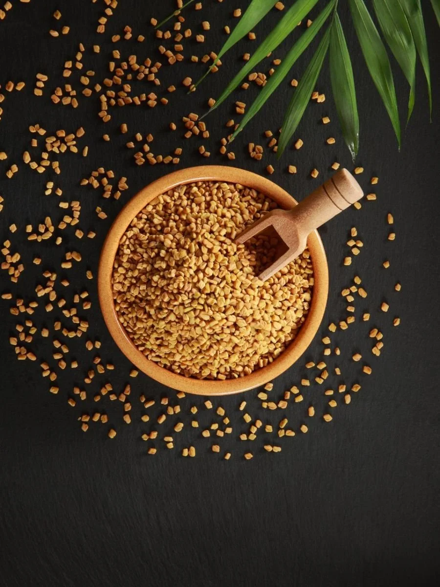 A wooden bowl filled with yellow split chickpeas on a black surface, with some chickpeas spilled around the bowl. A small wooden scoop rests inside the bowl. Green leaves are visible in the top right corner.