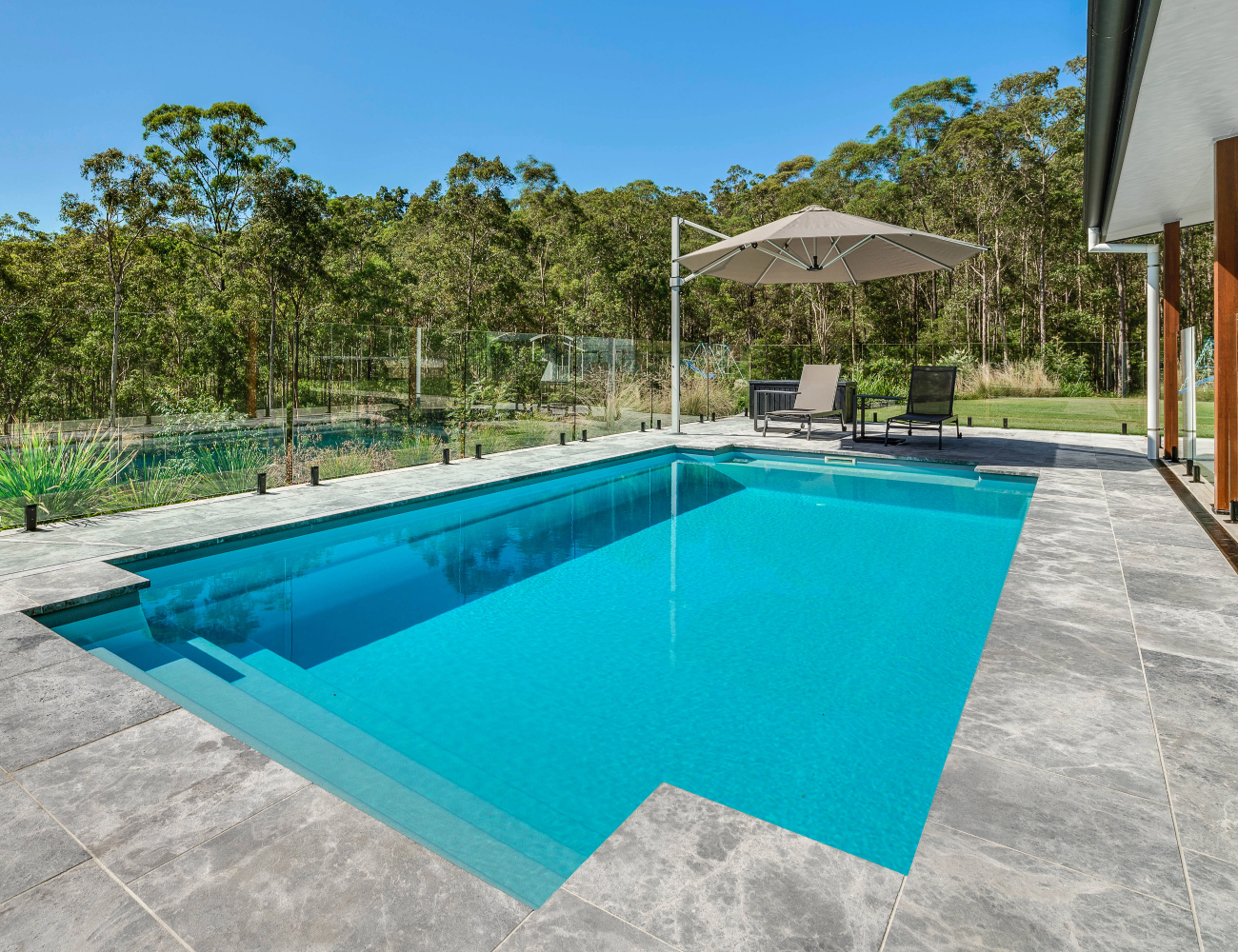 A modern backyard swimming pool with a gray tile deck, surrounded by glass fencing, with outdoor chairs and an umbrella, set against a backdrop of trees and a blue sky.