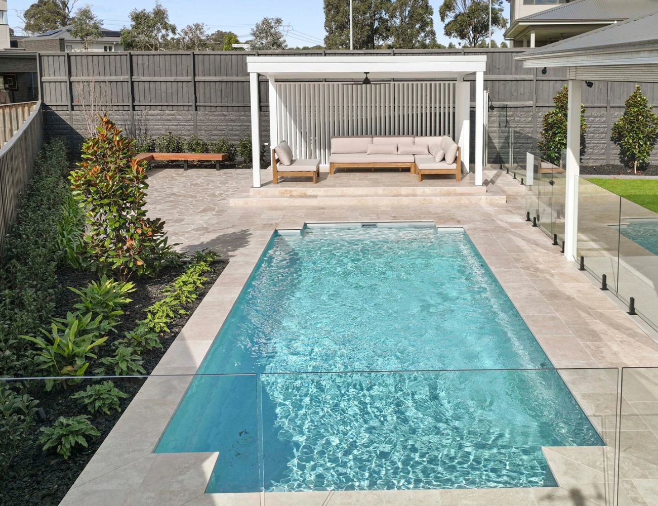 A backyard swimming pool surrounded by beige tiles, with a covered seating area that includes beige outdoor furniture and a small bench. The yard is enclosed with a wooden fence and has landscaped greenery along the sides.