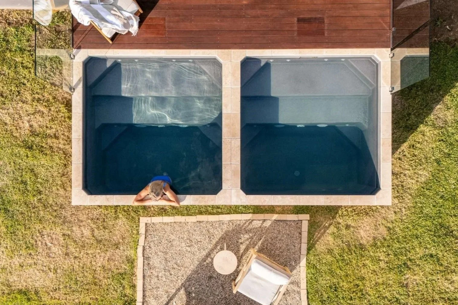 Overhead view of two side-by-side empty swimming pools with a woman leaning over the edge of the left pool. There is a wooden deck with chairs and a cover on the top side, and a small gravel area with a chair in the bottom part of the image.