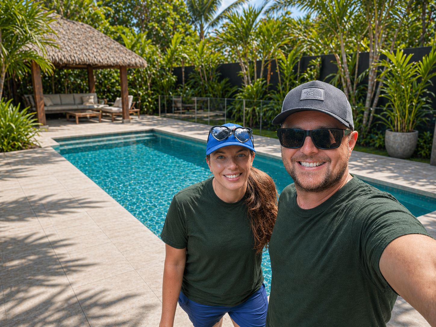 A smiling man and woman taking a selfie in front of a swimming pool, with lush greenery and a shaded lounge area in the background.