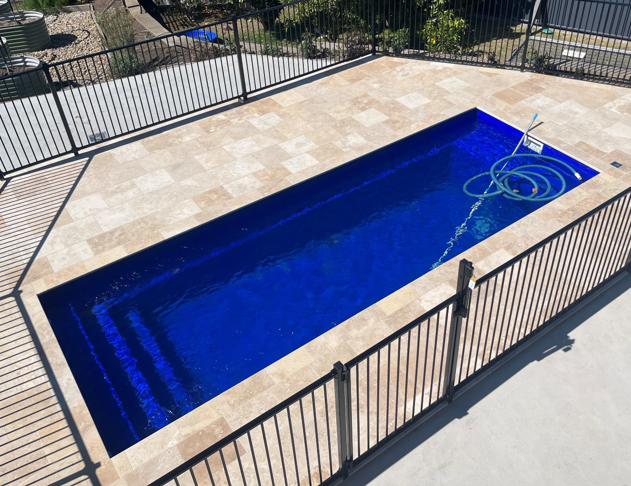 Empty rectangular backyard swimming pool with blue water, surrounded by a beige tiled deck and black metal fence.