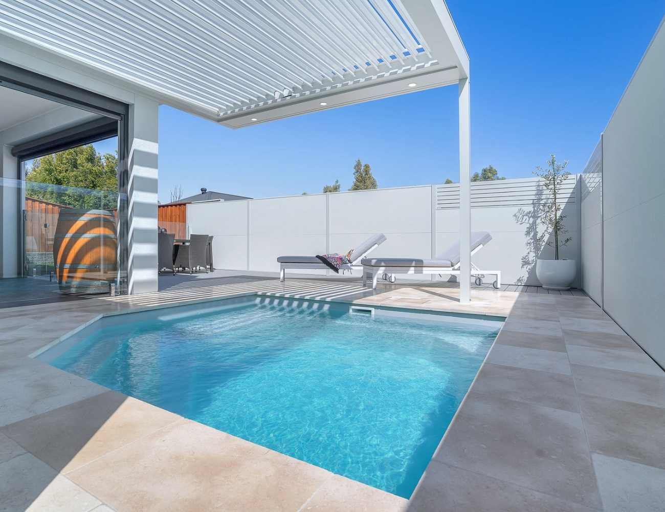 A modern backyard with a small swimming pool, two lounge chairs, a white fence, potted plants, and an outdoor dining area under a white pergola on a sunny day.