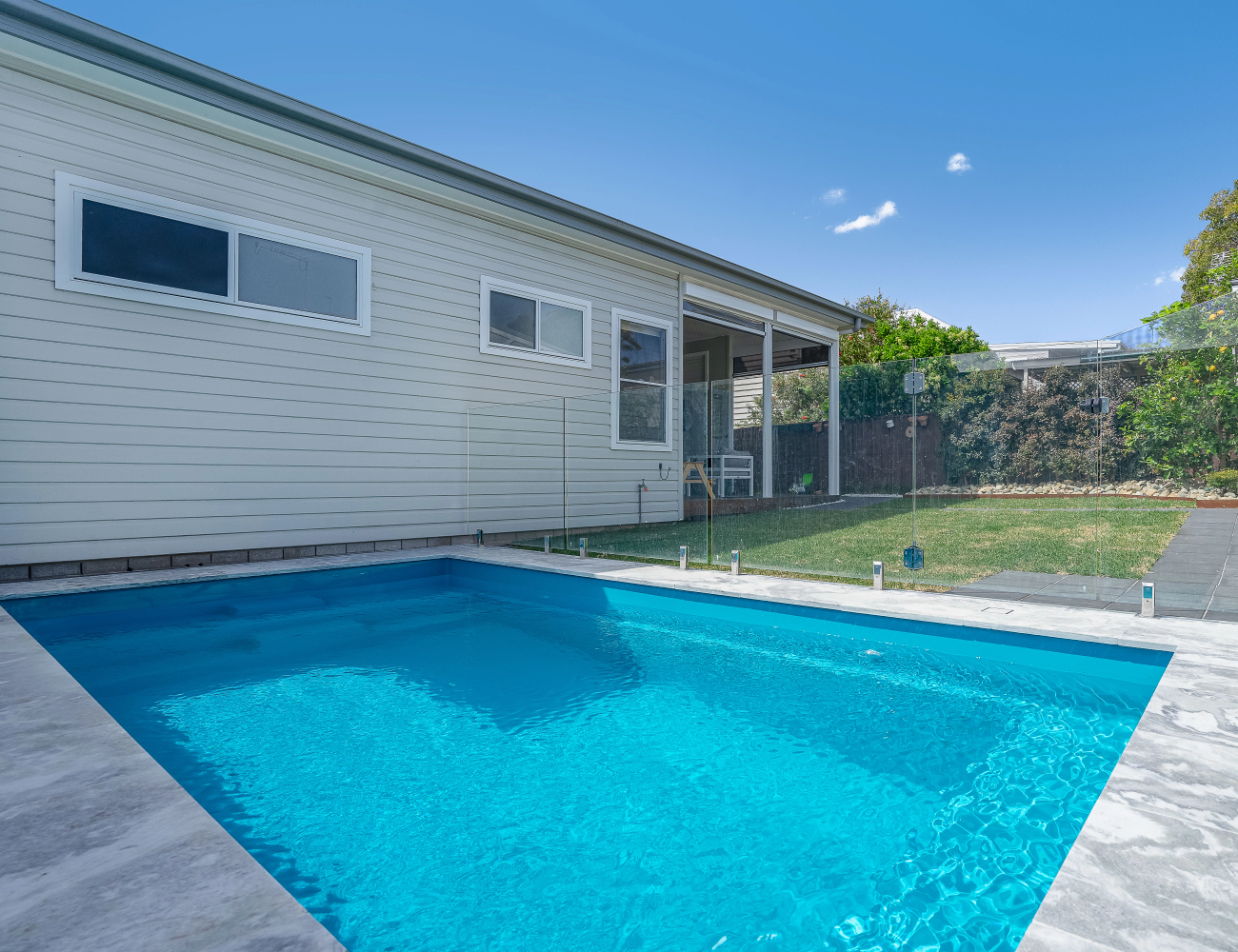 Backyard with a small swimming pool, white house siding, glass fencing, green grass, and trees under a blue sky.