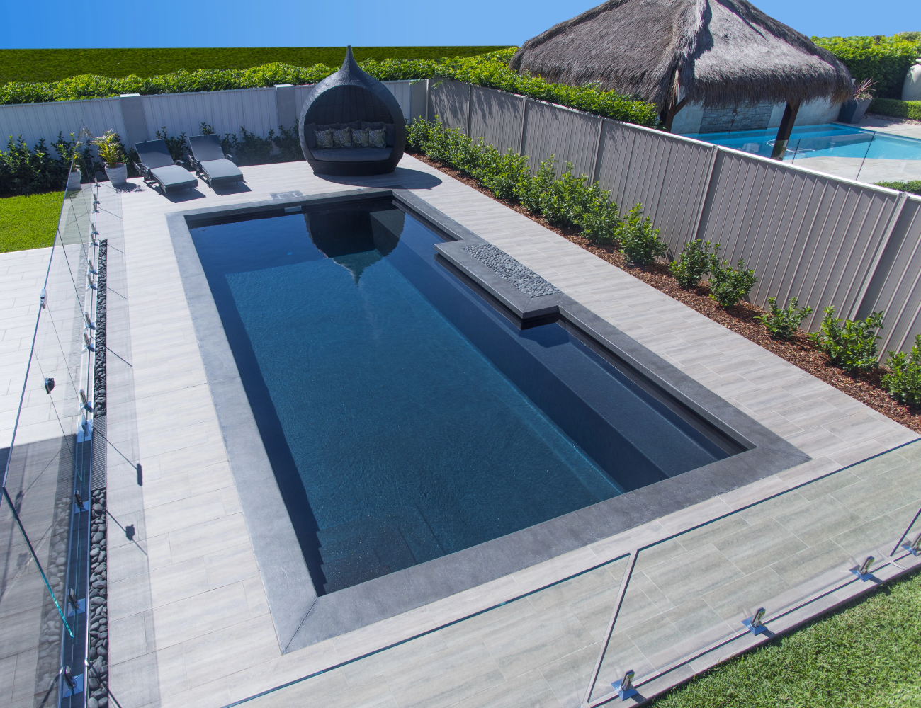 A backyard with a rectangular in-ground swimming pool, surrounded by a light gray patio, with a hot tub at the back and two lounge chairs with cushions, fenced in with glass panels, with grass and hedges along the fence, and a thatched-roof cabana visible in the background.