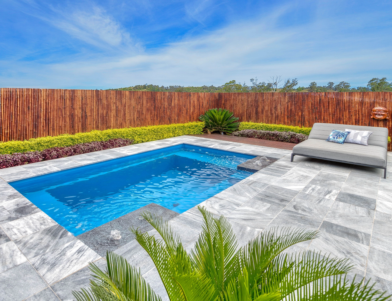 A backyard with a small swimming pool, surrounded by gray tile flooring and a wooden privacy fence. There is a gray lounge chair with pillows on it, some plants, and a clear blue sky.