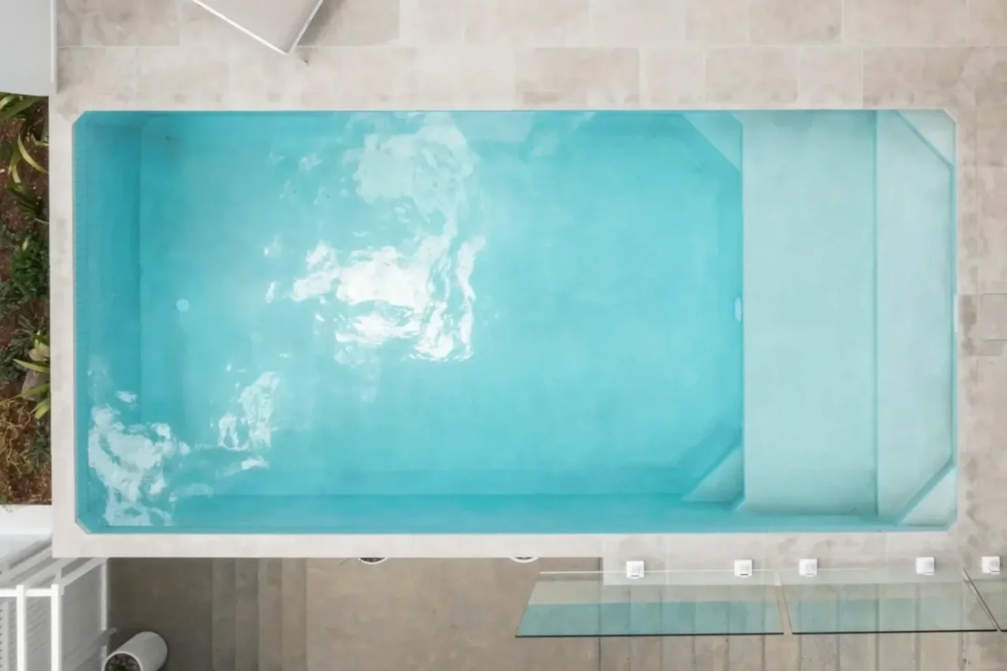 Overhead view of a rectangular swimming pool with light blue water, surrounded by light-colored tiled flooring with a small set of steps on one side.