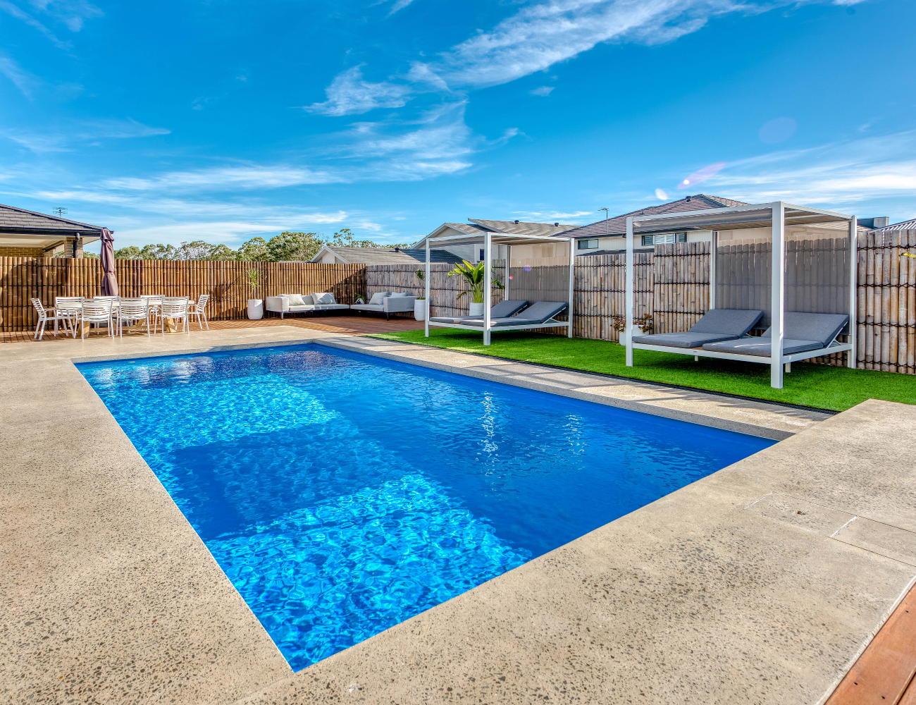 A backyard with a swimming pool, outdoor furniture, and cabana-style lounge chairs under a blue sky with clouds.