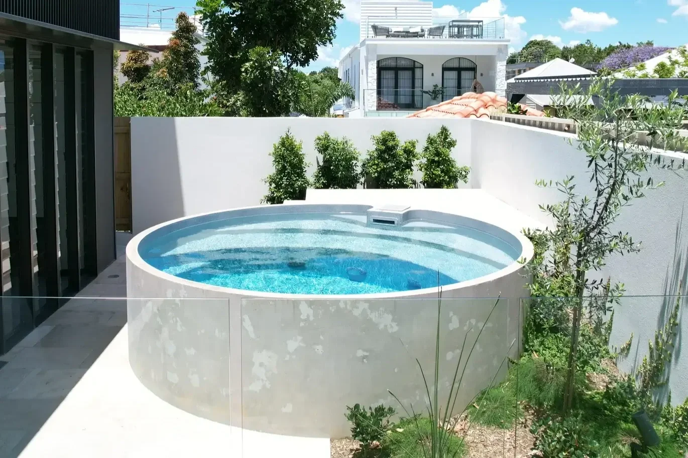 White-walled backyard with a round hot tub filled with water, surrounded by greenery and modern residential buildings in the background.