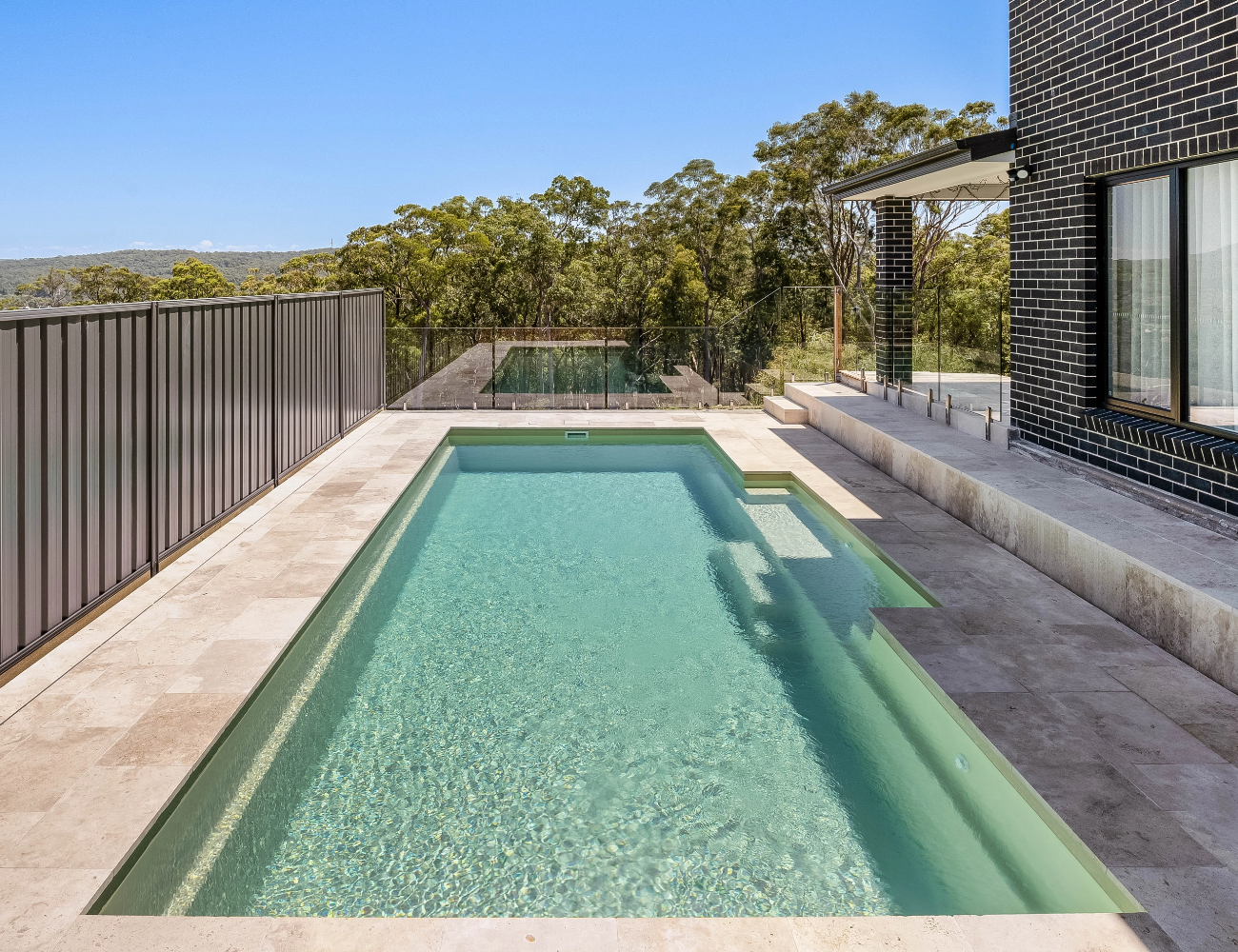 Modern backyard with a rectangular swimming pool, stone deck, black brick house, glass railing, and trees in the background.