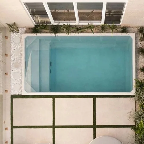 An aerial view of a modern backyard with a rectangular swimming pool, surrounded by patio tiles, green grass strips, and plants along the neighboring building.