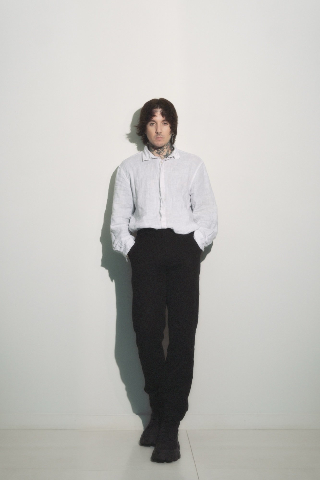 A person with dark curly hair and tattoos on the neck, wearing a white button-up shirt and black pants, standing against a plain white wall.