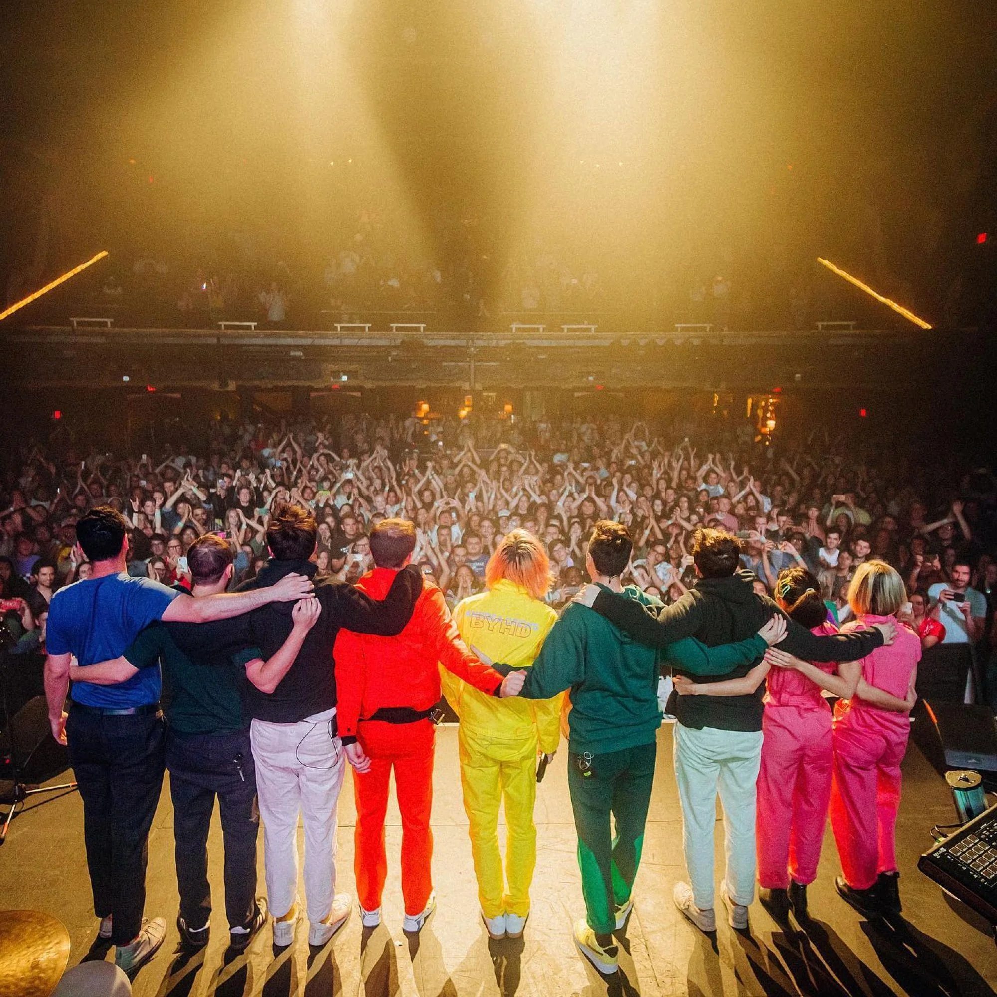 Group of people dressed in colorful tracksuits standing arm in arm at an indoor event with a large audience and stage lights in the background.