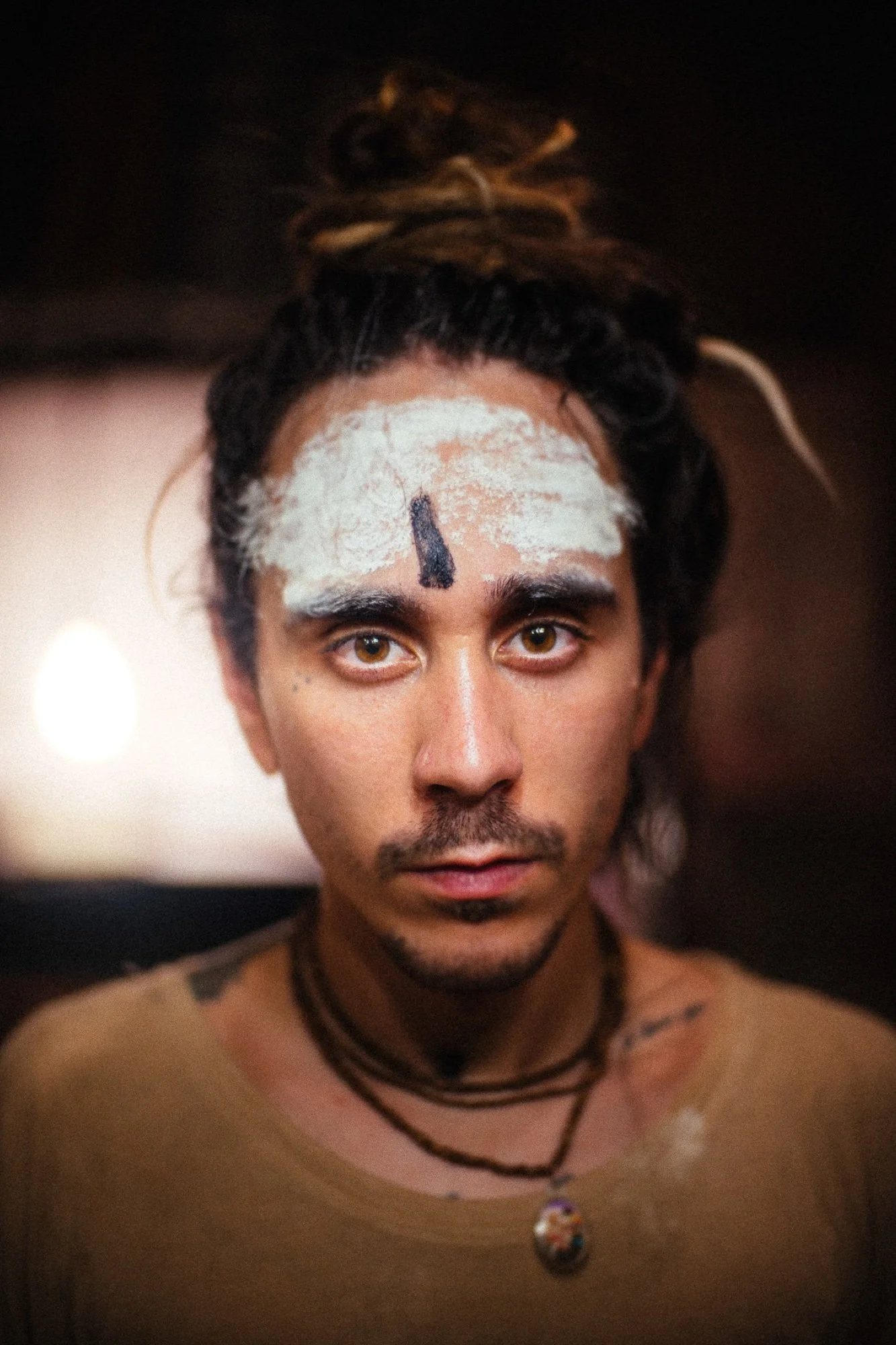 A person with a serious expression, face painted with white and black markings, has brown eyes, dark curly hair styled in a bun, wearing layered necklaces and a beige top, photo taken indoors with warm lighting.