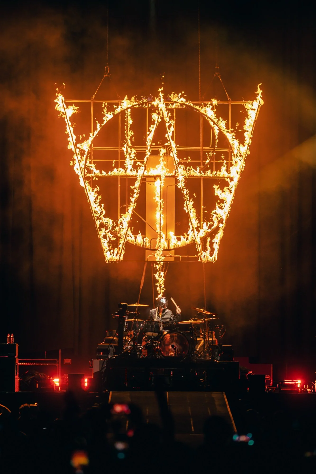 A music concert stage with a backdrop featuring the AVW logo engulfed in flames, and a drummer playing on a drum set in front of it.