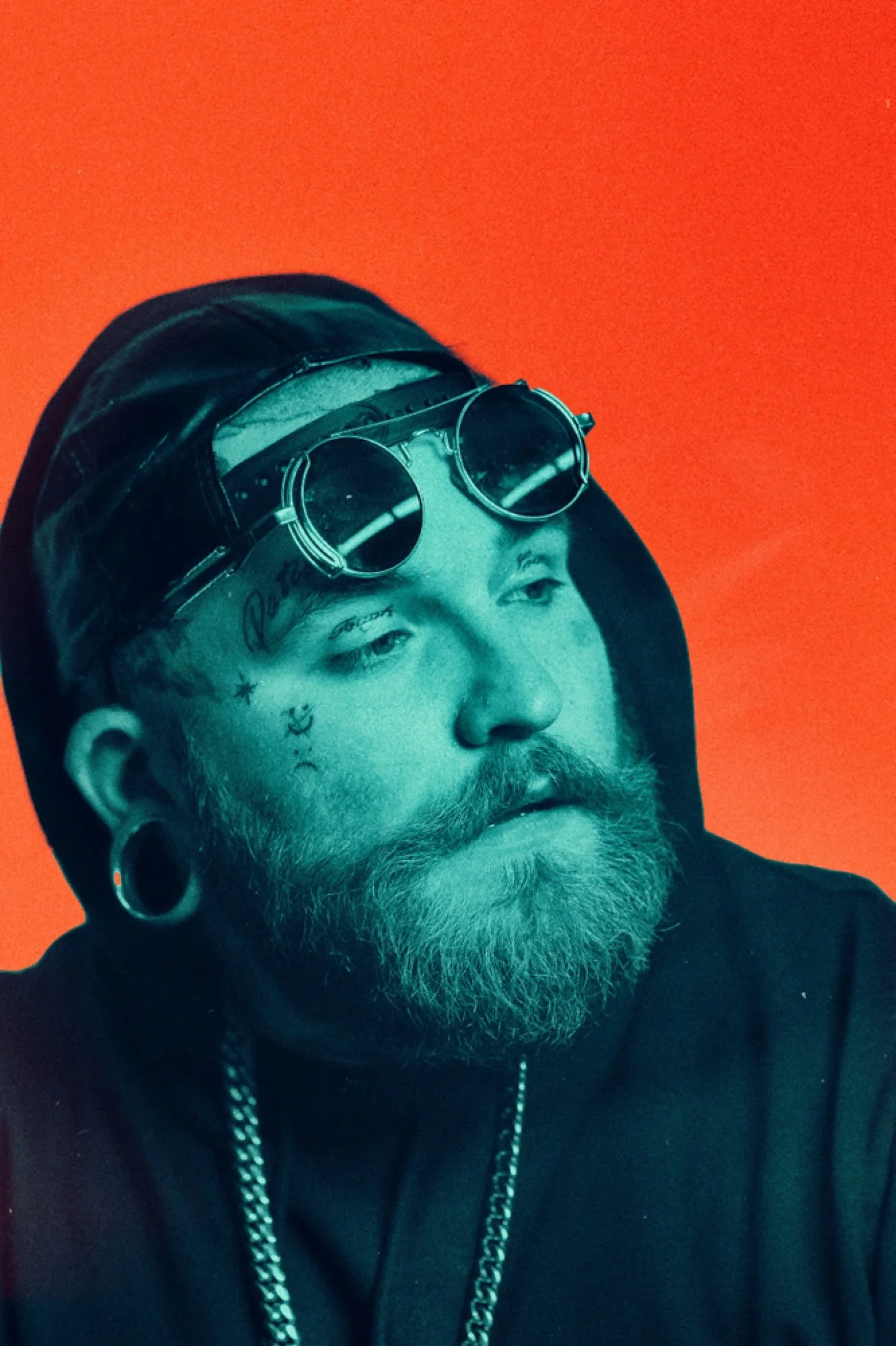 Close-up of a man with tattoos on his face and neck, wearing black sunglasses, a black hoodie, and a chain necklace, with an orange background.
