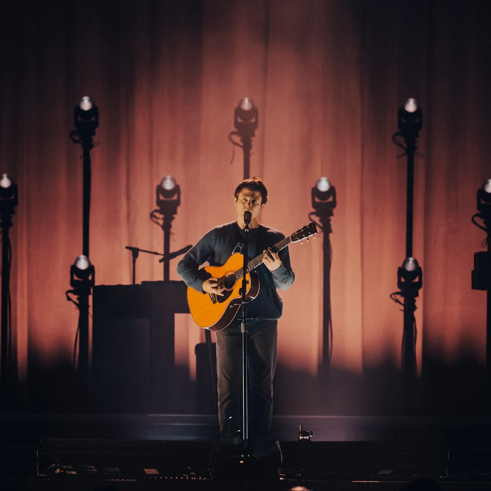 A man with short hair holding an acoustic guitar on stage, lit by stage lights with shadows cast on a curtain behind him.