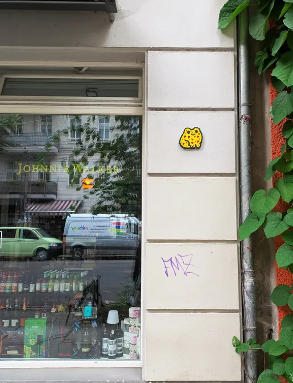 A storefront window with a green car and a van parked outside on the street. The window has the words 'JOHNNIE WALKER' in yellow lettering. Inside the store, bottles and products are visible. Adjacent to the window is a white wall with a yellow sticker of a leopard's face, and purple graffiti below it. Green leaves grow from the right side of the image.