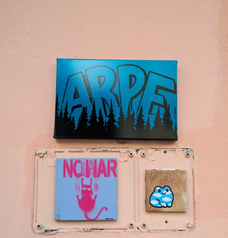 Three pieces of street art on a pink wall: a large blue and black sign with the word 'ARPE' and a forest silhouette, a pink and blue poster with a cartoon mouse and the word 'NOMAR,' and a small square piece with a cloud-like creature.
