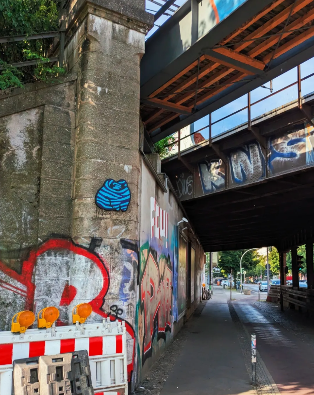Graffiti and street art on a concrete wall under a bridge, with construction barriers and orange warning lights in the foreground, and a sidewalk and street with trees and traffic in the background.