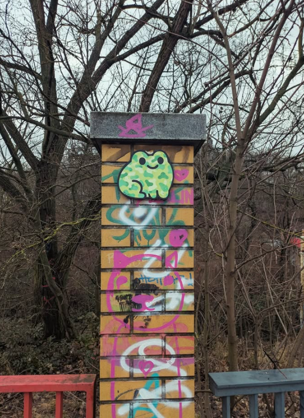 A small brick structure covered in colorful graffiti art, including a cute green creature, hearts, and various tags, with leafless trees in the background.