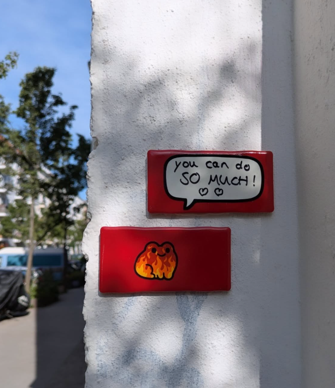 Two red rectangular signs on a white wall. The top sign has a speech bubble with handwritten text saying, 'you can do SO much!' with two small hearts. The bottom sign features a cute illustration of a fiery, orange, and yellow kitten.