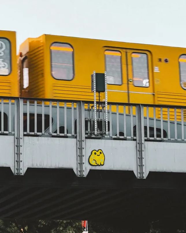 An elevated train on a bridge, with a small yellow cartoon cat sticker attached to the bridge rail. Yellow Frog attached to the wall.
