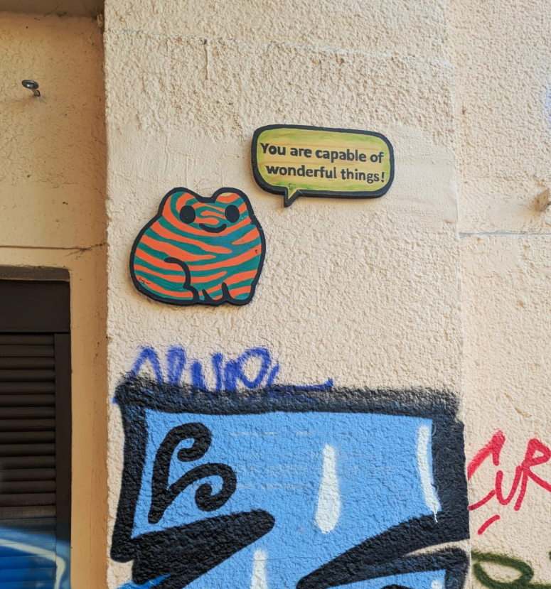 Colorful frog-shaped cartoon sign with a speech bubble that reads "You are capable of wonderful things!" on a textured beige wall with graffiti.