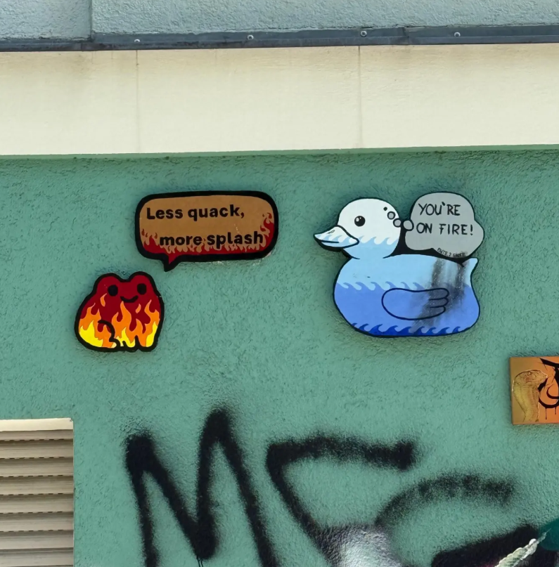 Graffiti art of a cartoon duck with a speech bubble saying "You're on fire!", a cloud with a face, and a fire character with a speech bubble saying "Less quack, more splash" on a green wall.