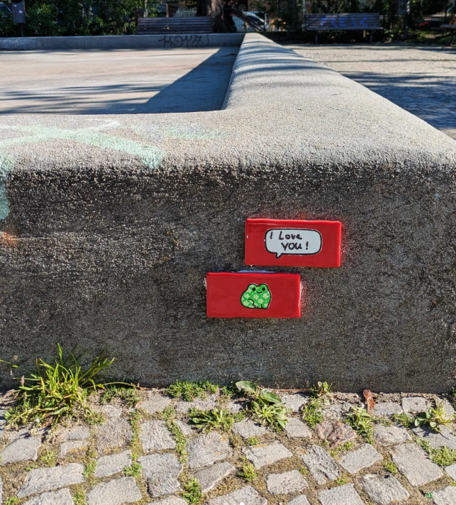 A concrete bench with two small red message plaques; one says "I love you!" and the other has a green frog illustration, located outdoors with grass and cobblestone pavement in the foreground.