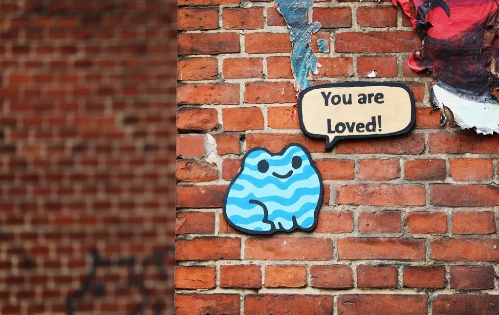 A decorative brick wall with a cartoon-style blue creature with black eyes and a smiling face, accompanied by a speech bubble that says 'You are Loved!' and torn pieces of paper above.