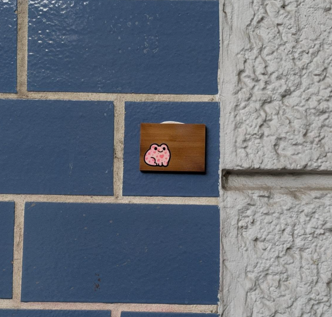 A small pink sticker of a cartoon-style animal, possibly a hippo, on a brown wooden block attached to a blue brick wall.